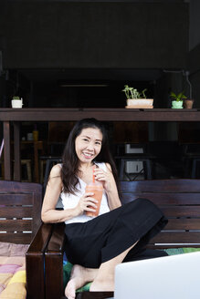 Portrait of smiling woman sitting on terrace bench drinking a smoothie - IGGF00207