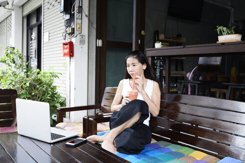 Woman sitting on terrace bench drinking a smoothie and looking at laptop - IGGF00208