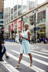 USA, New York, junge blonde afro-amerikanische Frau mit Tasse Kaffee und Smartphone beim Überqueren der Straße - MAUF01238