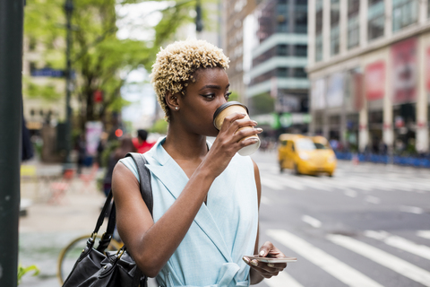 USA, New York, junge blonde afro-amerikanische Frau mit Kaffeetasse und Smartphone, lizenzfreies Stockfoto
