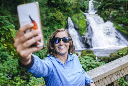Germany, Triberg, woman taking a selfie with mobile phone in front of Triberg Waterfalls - KIJF01720