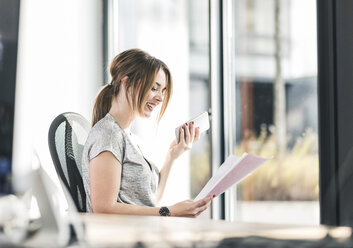 Smiling businesswoman with cell phone and documents at desk in office - UUF12404