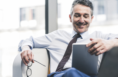 Lächelnder Geschäftsmann sitzt mit Tablet im Wohnzimmer, lizenzfreies Stockfoto