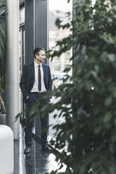 Businessman standing in office lobby looking out of window - UUF12457