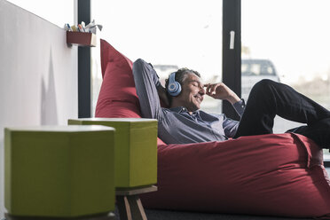 Smiling businessman sitting in beanbag wearing headphones - UUF12465