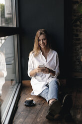 Young woman sitting on window sill in a cafe, using smartphone - GUSF00199