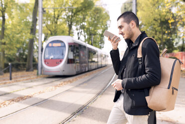 Junger Mann mit Mobiltelefon an der Straßenbahnhaltestelle im Freien - FMOF00324