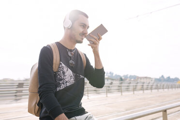 Junger Mann mit Kopfhörern und Mobiltelefon an einer Straßenbahnhaltestelle im Freien - FMOF00329