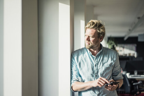 Geschäftsmann mit Mobiltelefon im Büro und Blick aus dem Fenster - JOSF02025