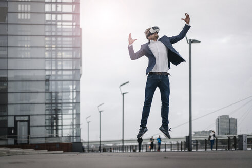 Businessman wearing VR glasses jumping in the city - JOSF02058