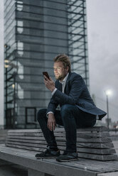 Geschäftsmann sitzt auf einer Bank in der Stadt und hält ein Mobiltelefon - JOSF02072