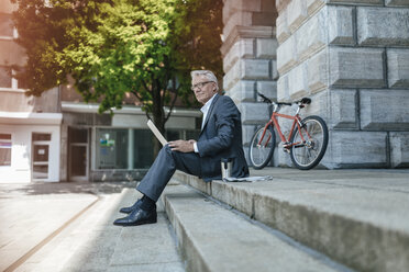 Senior businessman sitting on stairs, using laptop - GUSF00256