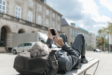 Senior businessman using smartphone, lying on bench in the city - GUSF00260