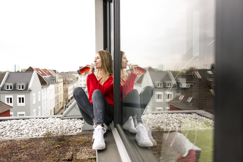 Lächelnde junge Frau, die in einer Stadtwohnung am Fenster sitzt und einen Drink zu sich nimmt, lizenzfreies Stockfoto