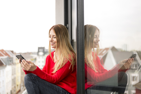Smiling young woman sitting at the window in city apartment looking at cell phone stock photo