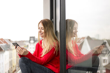 Lächelnde junge Frau sitzt am Fenster in einer Stadtwohnung und schaut auf ihr Handy - PESF00804