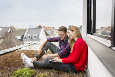 Smiling young couple sitting on roof of city apartment using laptop - PESF00808