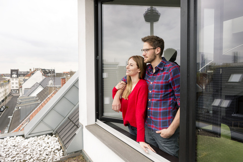 Lächelndes junges Paar schaut aus dem Fenster in einer Stadtwohnung, lizenzfreies Stockfoto