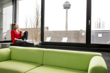 Smiling young woman sitting at the window in city apartment - PESF00810