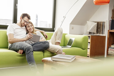 Smiling young couple on couch in living room at home sharing tablet - PESF00832
