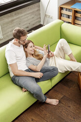 Smiling young couple on couch in living room at home sharing cell phone - PESF00835