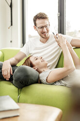 Smiling young couple on couch in living room at home sharing cell phone - PESF00837