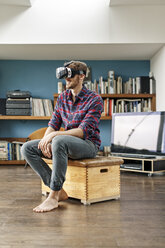 Junger Mann zu Hause auf einer Kiste sitzend mit VR-Brille - PESF00839