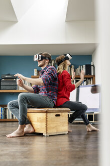 Junges Paar zu Hause, das auf einer Kiste sitzt und eine VR-Brille trägt - PESF00840