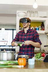 Happy young man wearing VR glasses cooking in kitchen - PESF00853
