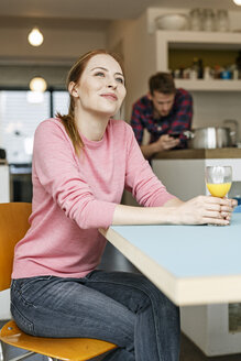 Lächelnde junge Frau mit einem Glas Orangensaft in der Küche zu Hause mit einem Mann im Hintergrund - PESF00856