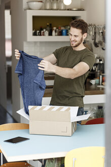 Lächelnder junger Mann zu Hause beim Auspacken eines Pakets mit Kleidung - PESF00867