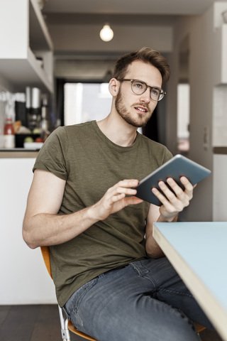 Junger Mann mit Tablet sitzt in der Küche zu Hause, lizenzfreies Stockfoto