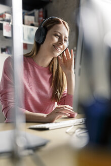 Smiling young woman wearing headphones at desk at home - PESF00873