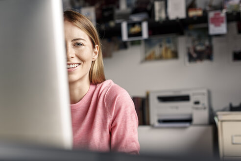 Porträt einer lächelnden jungen Frau hinter einem Computerbildschirm am Schreibtisch zu Hause - PESF00882