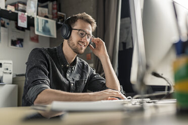 Lächelnder junger Mann mit Kopfhörern, der am Schreibtisch zu Hause am Computer arbeitet - PESF00885