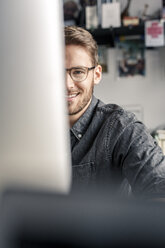 Portrait of smiling young man behind computer screen at desk at home - PESF00887