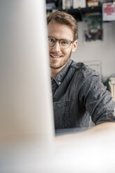 Portrait of smiling young man behind computer screen at desk at home - PESF00888