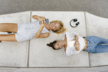 Two happy young women with cell phones lying on ramp in a skatepark - KNSF03065