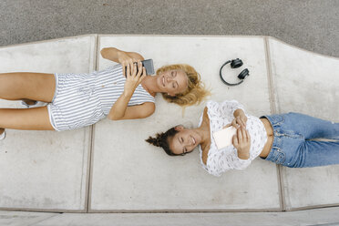 Zwei glückliche junge Frauen mit Handys auf einer Rampe in einem Skatepark liegend - KNSF03065