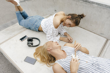 Zwei glückliche junge Frauen liegen auf einer Rampe in einem Skatepark und machen Musik - KNSF03069