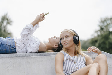 Zwei junge Frauen mit Handy und Kopfhörern in einem Skatepark - KNSF03110