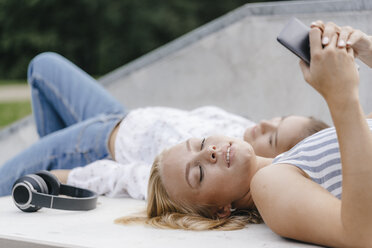 Zwei junge Frauen mit Handy und Kopfhörern liegen auf einer Rampe in einem Skatepark - KNSF03115