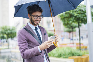 Modischer Geschäftsmann mit Regenschirm überprüft sein Handy in der Stadt - BSZF00098