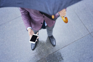 Geschäftsmann steht unter einem Regenschirm und hält ein Mobiltelefon, Teilansicht - BSZF00100