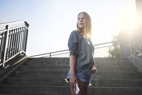 Blonde junge Frau mit Smartphone bei Gegenlicht vor einer Treppe stehend, lizenzfreies Stockfoto