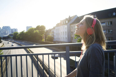 Lächelnde junge Frau auf einer Brücke, die mit Kopfhörern Musik hört - KNSF03148