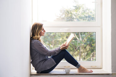 Woman sitting at home on the window sill, reading a book - HAPF02485