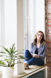 Woman sitting at home on the window sill, drinking coffee - HAPF02490