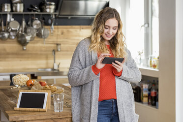 Portrait of smiling young woman using smartphone in the kitchen - FMKF04652