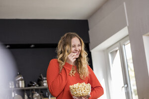 Porträt einer kichernden jungen Frau mit einer Schüssel Popcorn in der Küche - FMKF04653